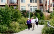 Older adults walk together on a scenic path near the Aljoya Thornton Place retirement community in Seattle, surrounded by greenery.