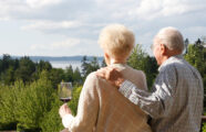 Older couple enjoying wine on a balcony at the Aljoya Mercer Island retirement community in Mercer Island, with a scenic view of the ocean and trees.