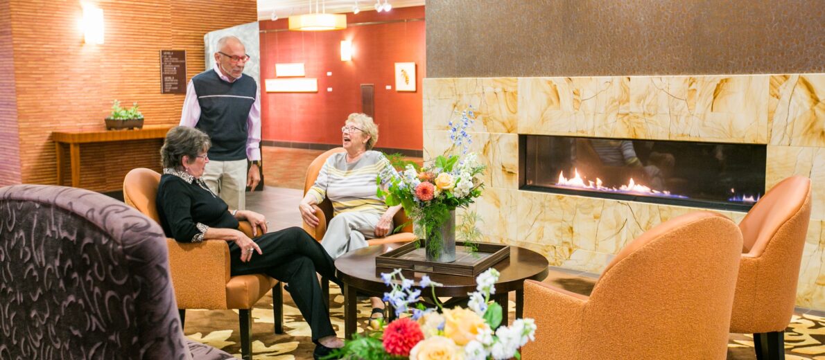 Older adults enjoying community and warmth by the fireplace in the lobby of the Aljoya Thornton Place retirement community in Seattle.