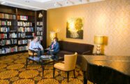 An older couple discusses literature in the inviting library at the Aljoya Thornton Place retirement community in Seattle, with comfortable seating and a piano.