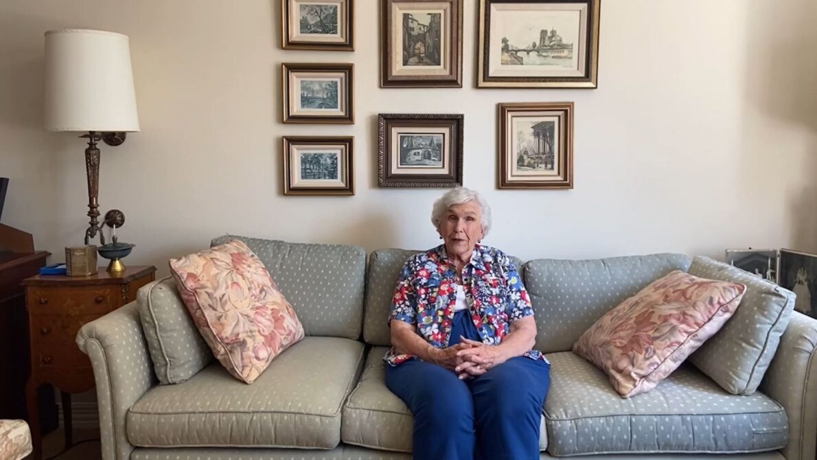 An older woman relaxes on a couch, with various family photos displayed on the wall behind her, evoking cherished memories at The Gardens at Town Square.