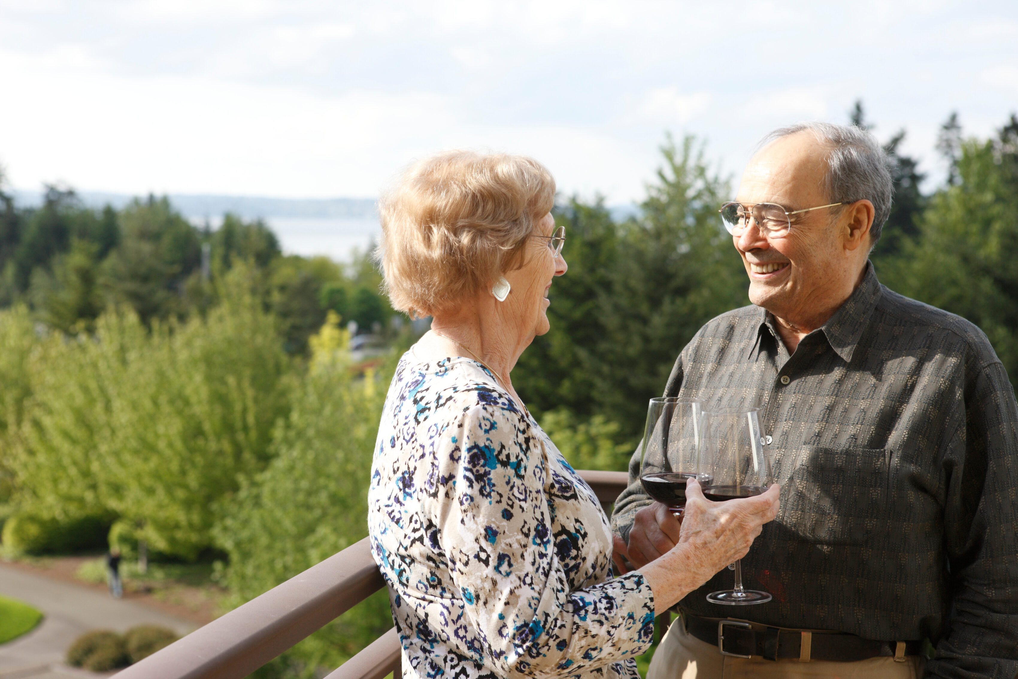 Older couple enjoying wine on a balcony at the Aljoya Mercer Island retirement community in Mercer Island, with a scenic view of the ocean and trees.