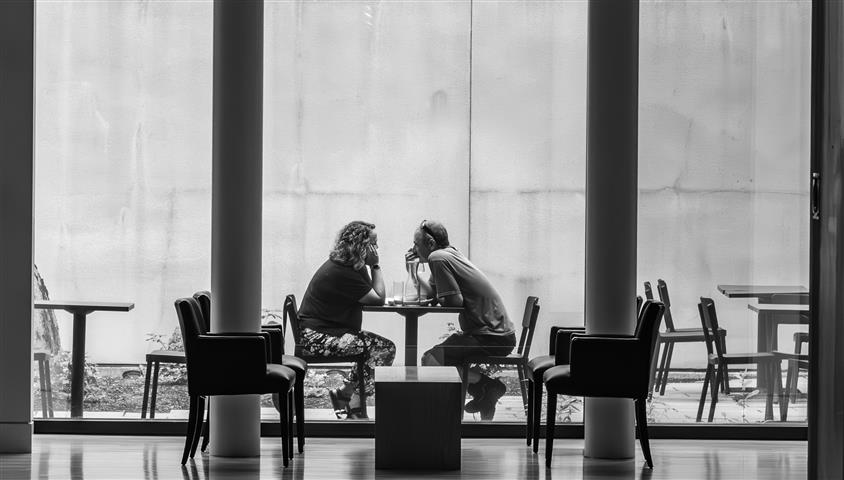Black and white photo of two people having a conversation in front of a large window.